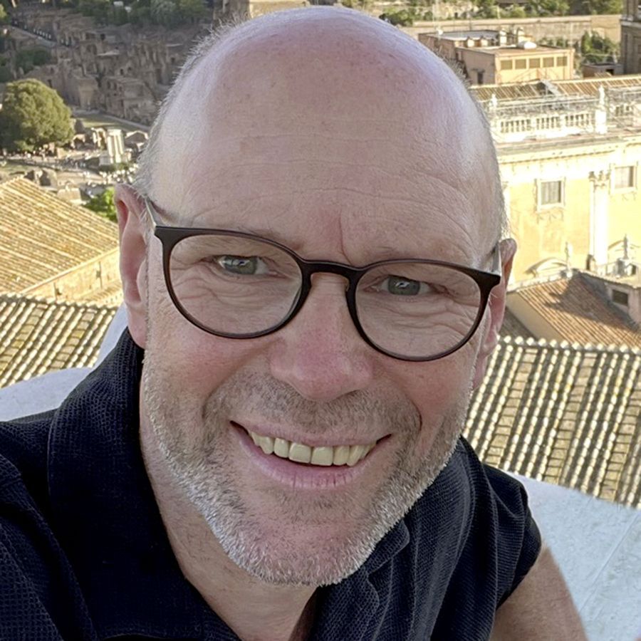 Glücklicher Mann mit Brille lächelt vor historischer Architektur im Hintergrund.
